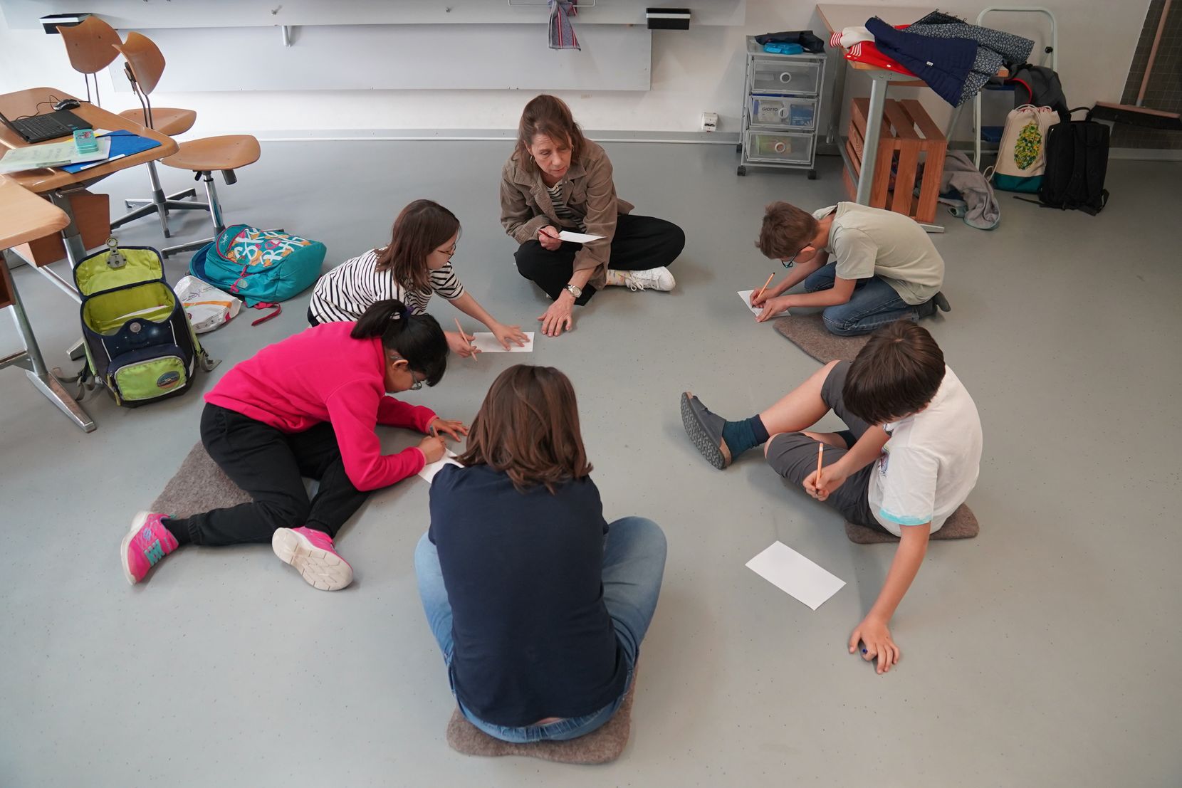 Kinder und eine Lehrerin sitzen im Kreis auf dem Boden eines Klassenzimmers.